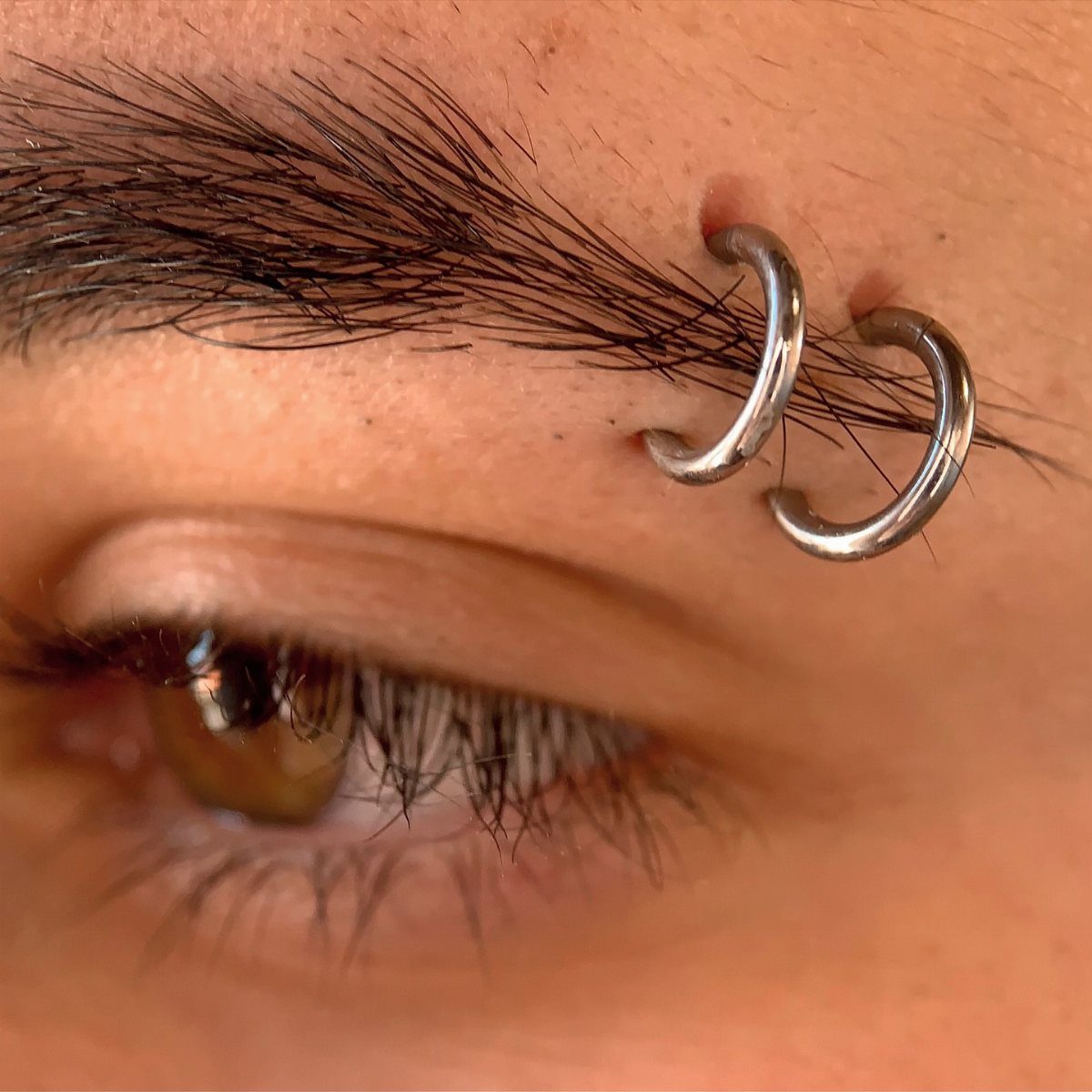 Two silver hoop piercings in a person's eyebrow above a brown eye.