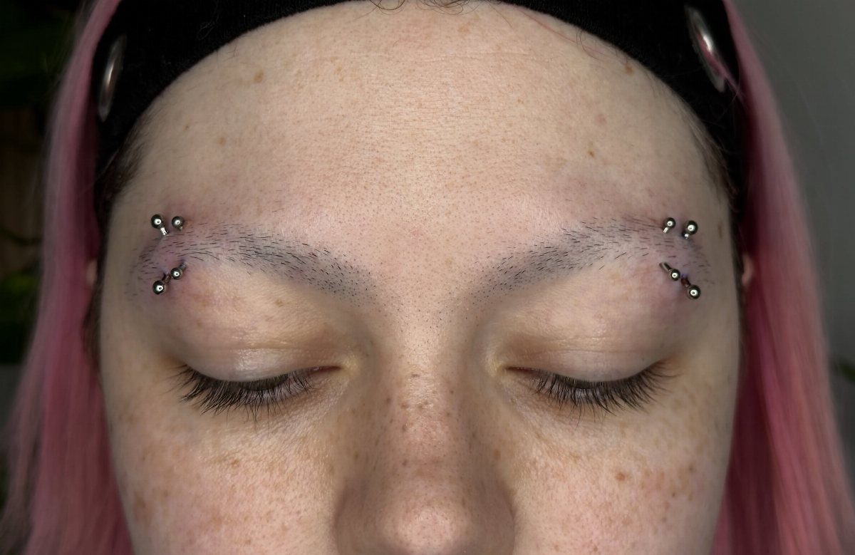 Person with pink hair showing double eyebrow piercings above and below each brow.