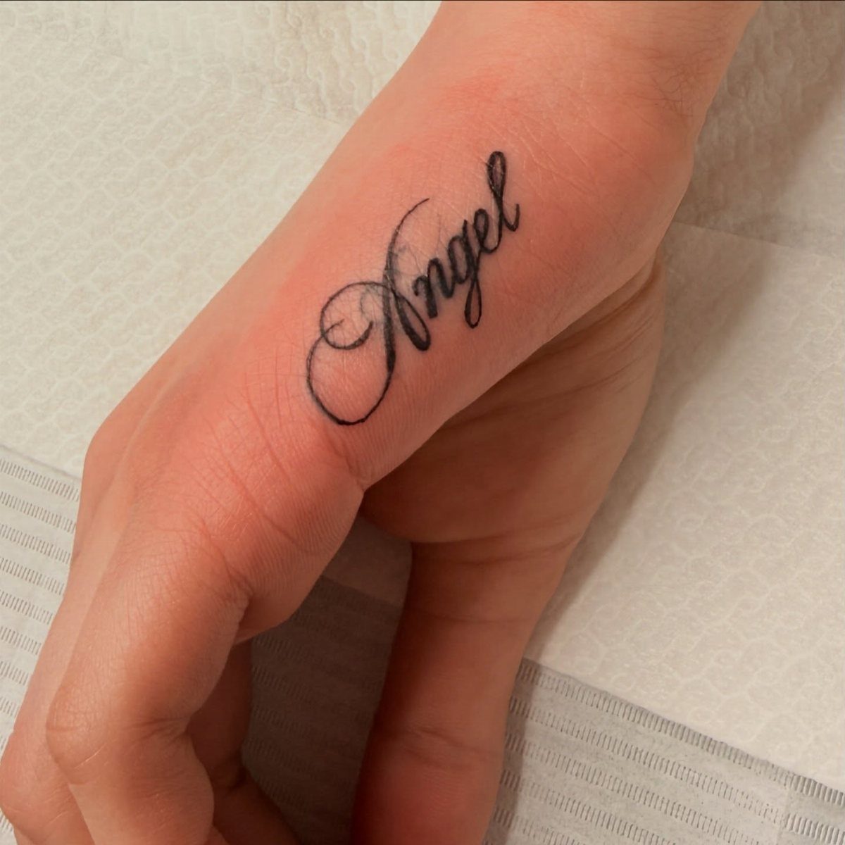 Cursive black ink tattoo of the word Angel on a person's hand.