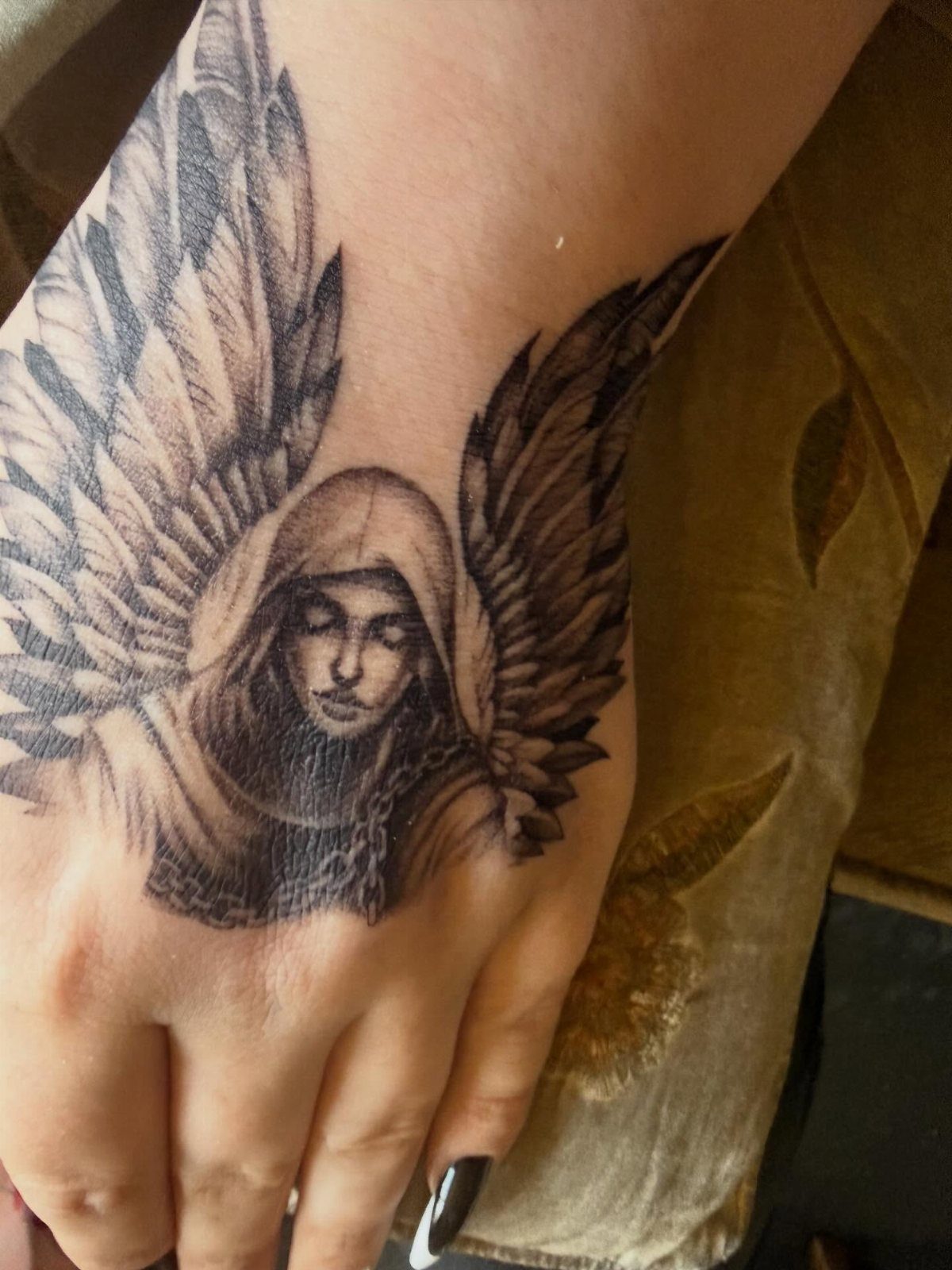 Hooded angel tattoo with large wings on the back of a hand.