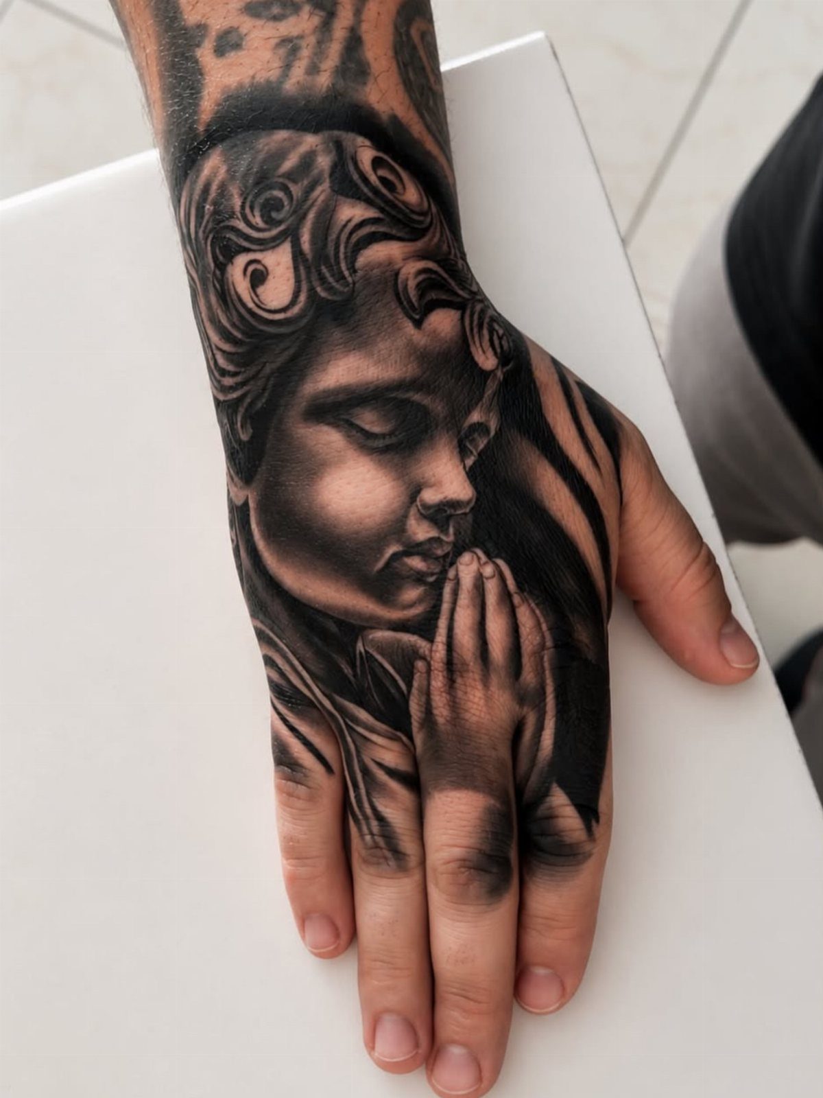 Praying cherub tattoo on a hand with detailed black and grey shading.