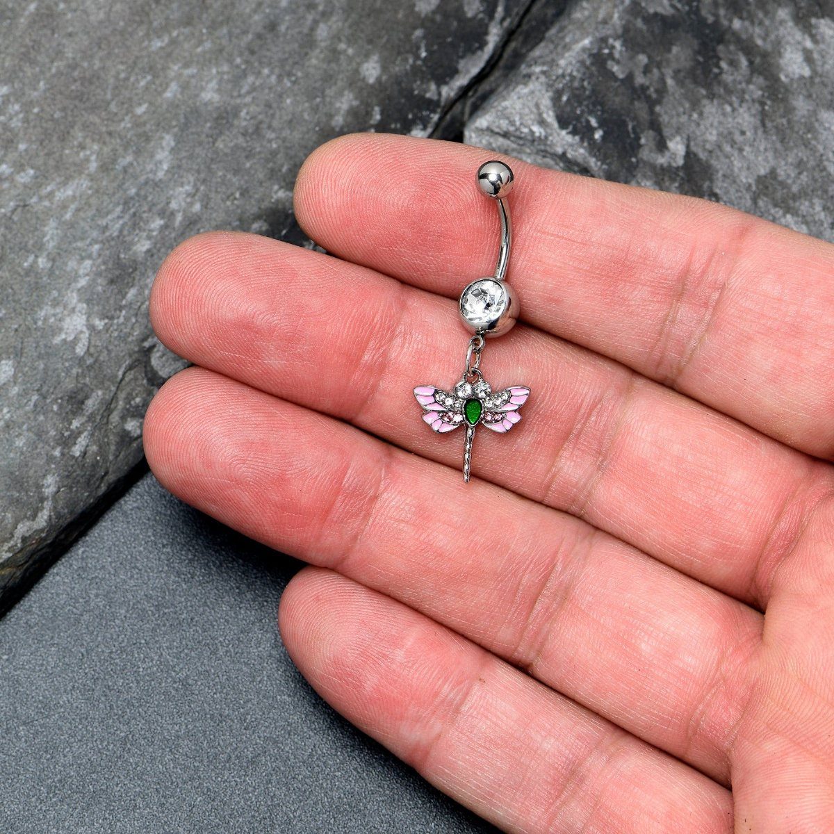 Silver-tone belly piercing barbell with a dangling pink crystal dragonfly charm held in hand.