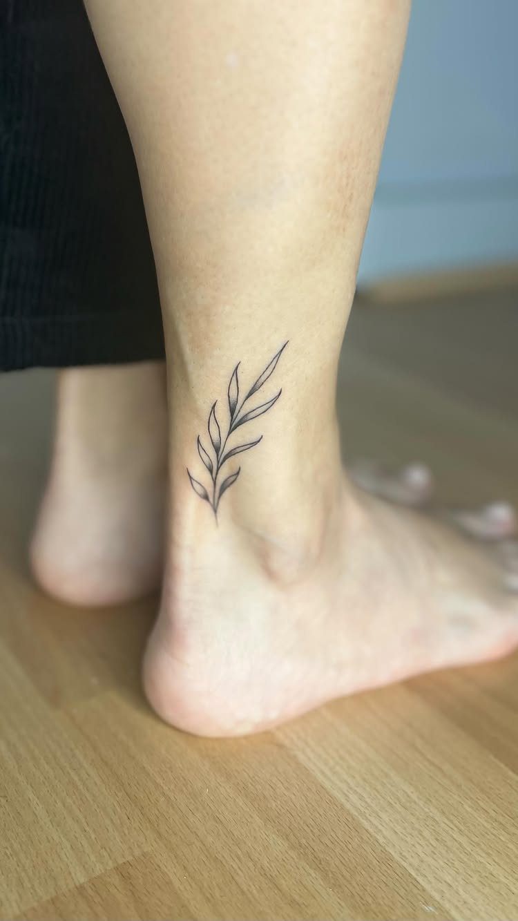Ankle tattoo of a delicate black leafy branch on a person's leg.