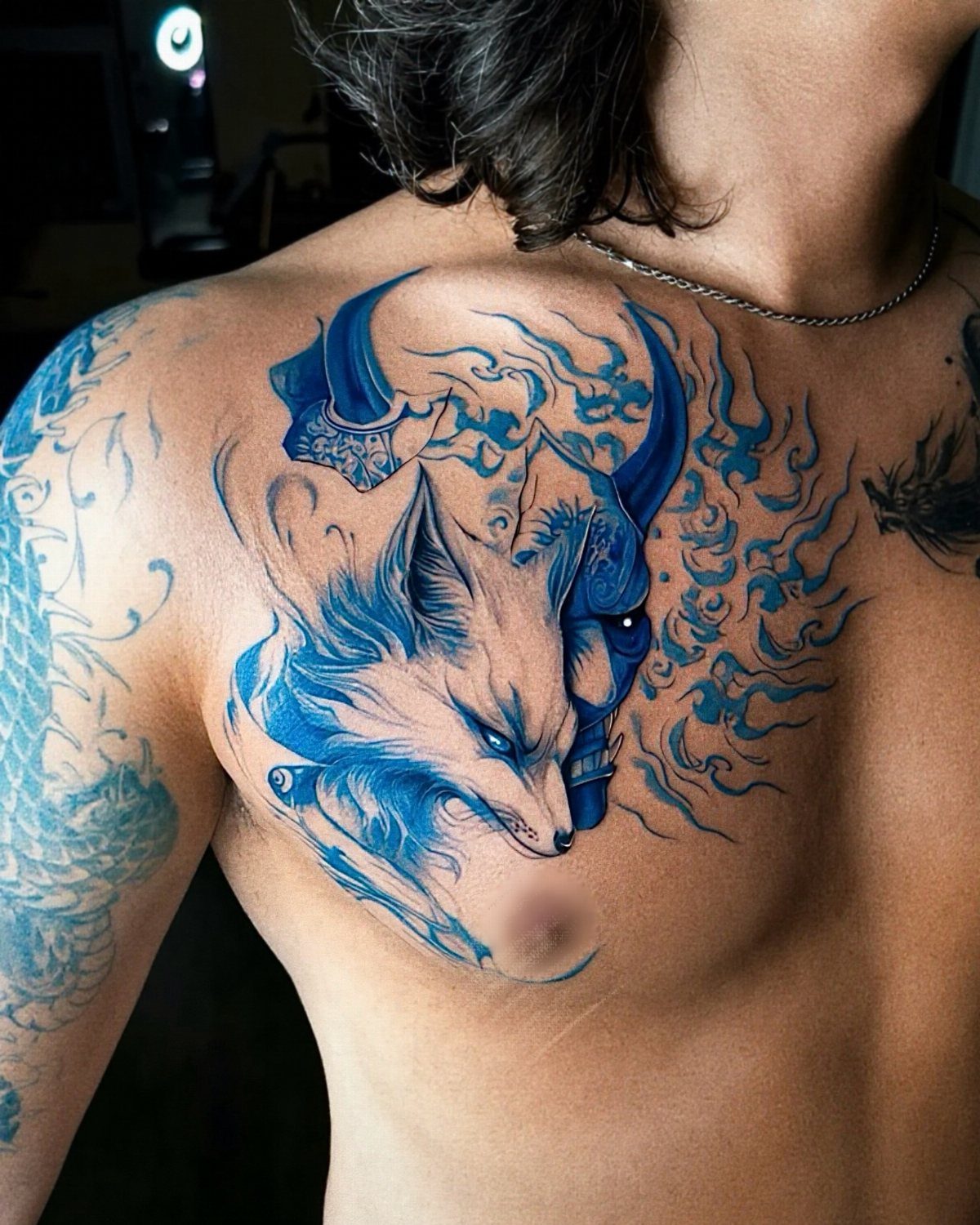 Blue fox and horned mask tattoo on a person's chest and shoulder.