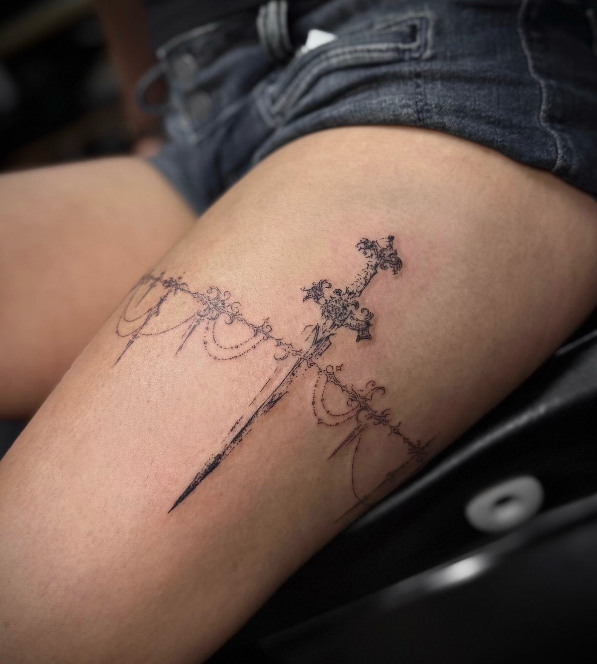 Black ink sword and filigree tattoo on a person's upper thigh.