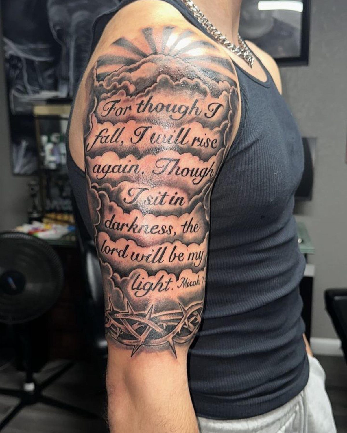 Upper arm tattoo with clouds, sun rays, and a religious quote in grey ink.