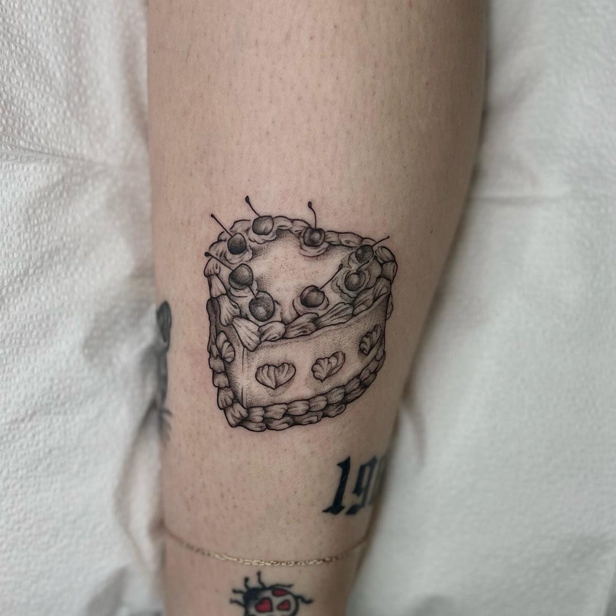Heart-shaped birthday cake tattoo with cherries on a person's arm.
