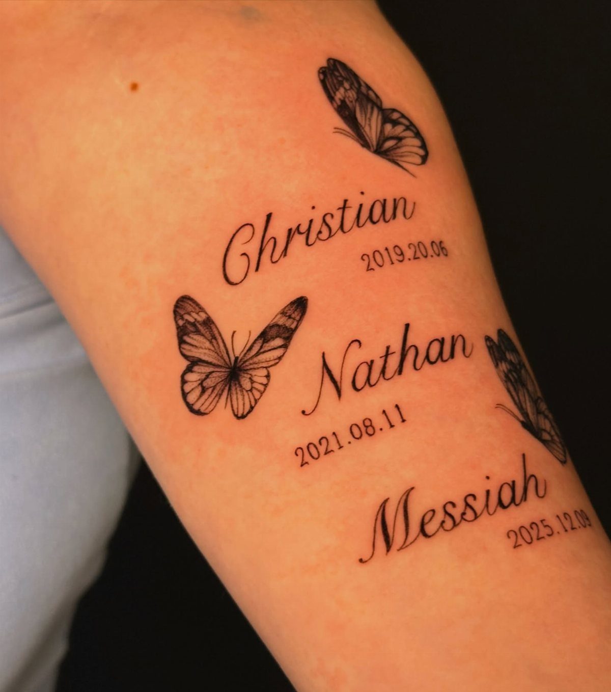 Tattoos of names and dates with butterflies on a person's forearm.