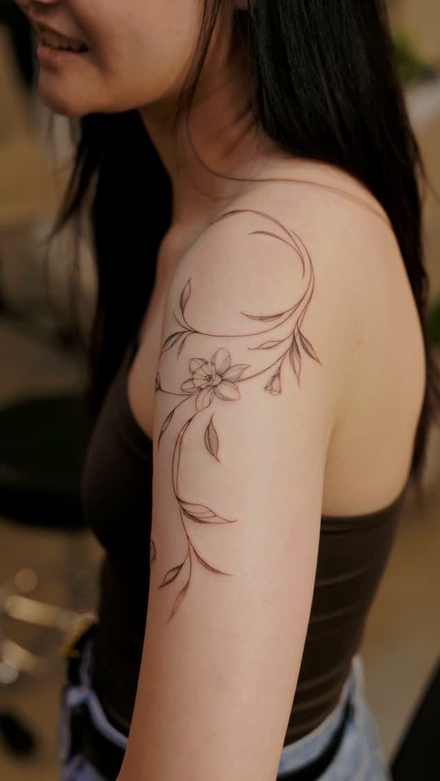Fine-line floral tattoo with a single flower and swirling vines on an upper arm.