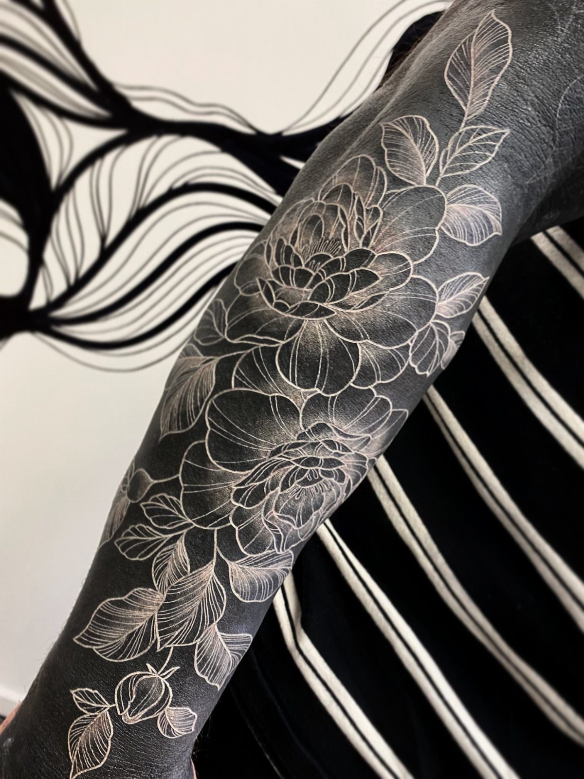White ink floral tattoo of peonies and leaves flowing vertically down an arm.