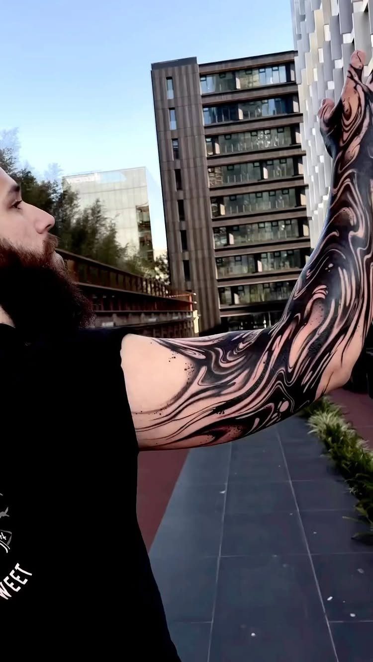 Man's arm with a swirling black ink sleeve tattoo against city buildings.