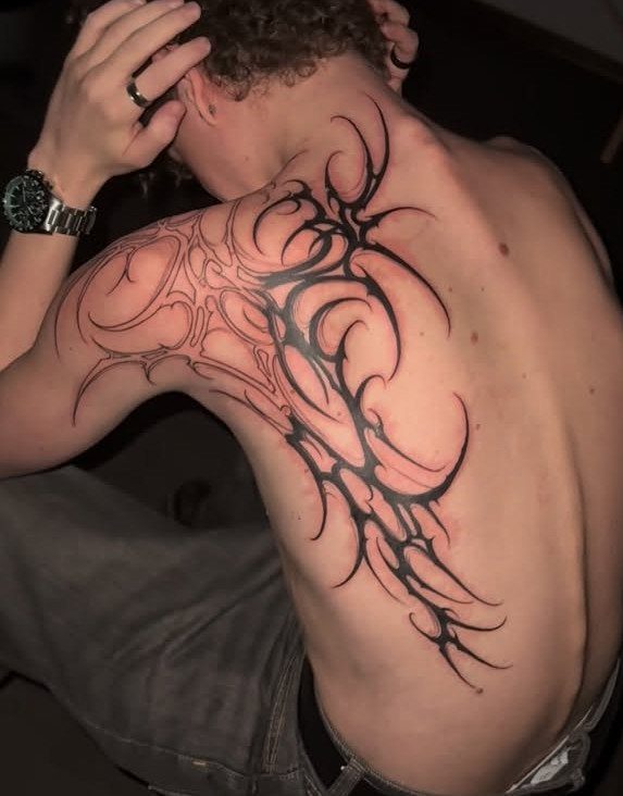 Tribal blackwork tattoo with sharp curving lines on a person's upper back and shoulder.