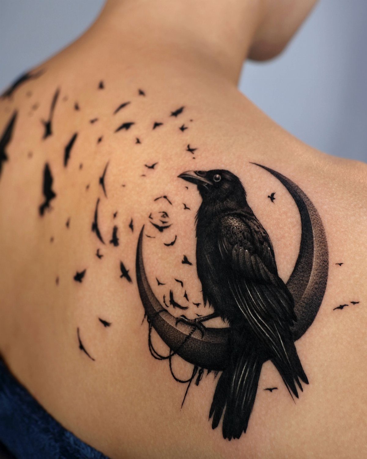 Blackbird perched on a crescent moon tattoo on a person's shoulder with flying birds.