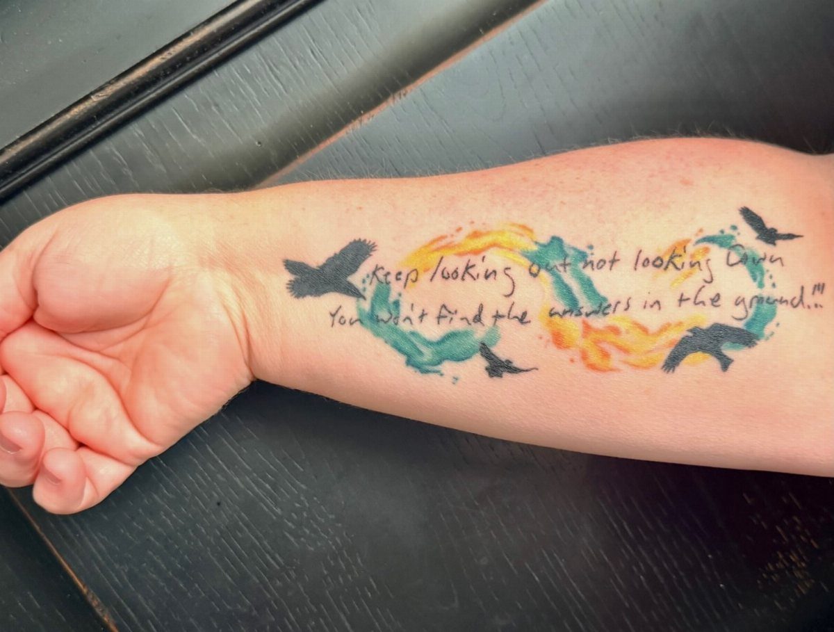 Tattoo on a forearm with black birds, a quote, and colorful watercolor splashes.