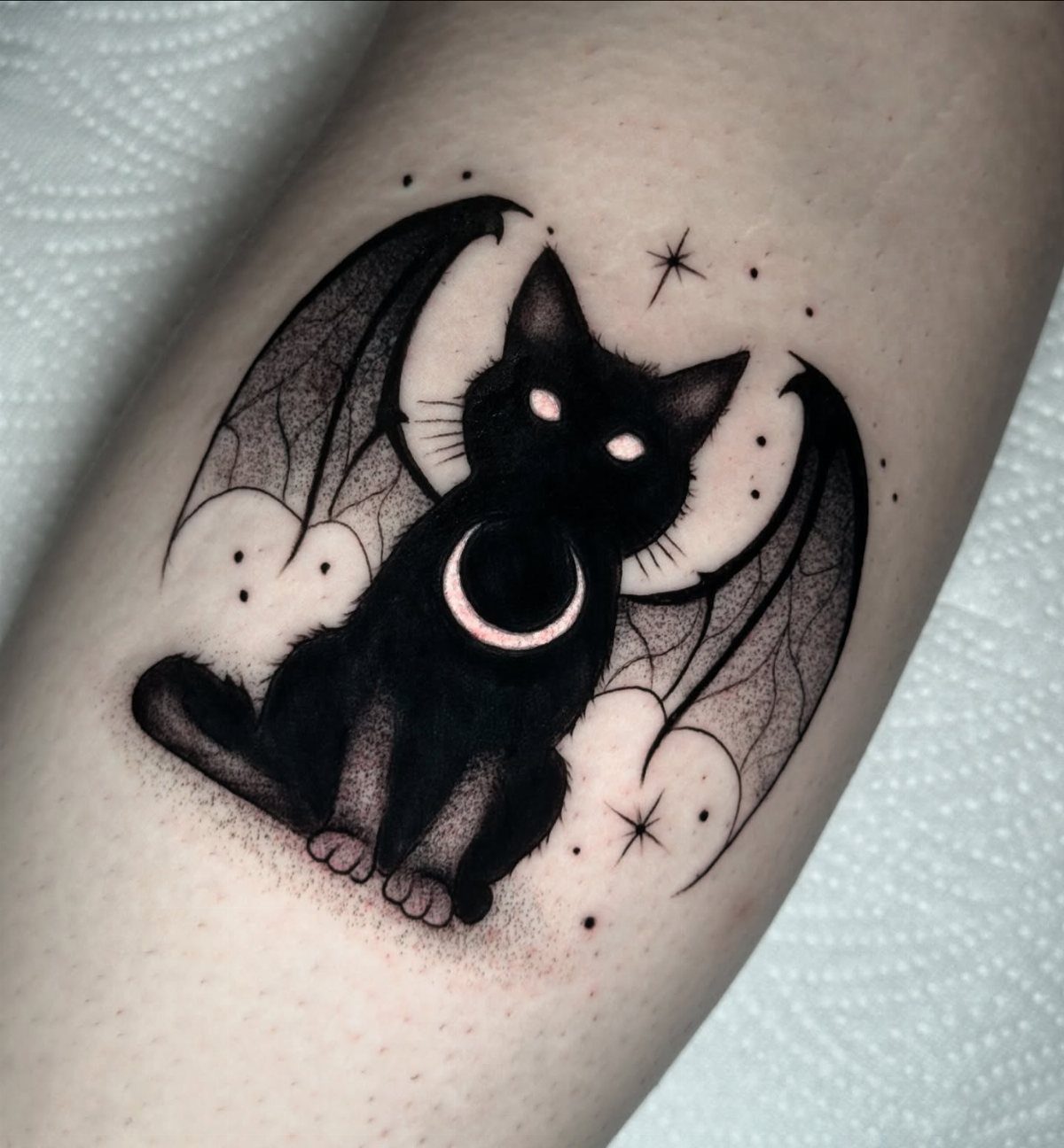 Black cat tattoo with bat wings and a crescent moon on human skin.