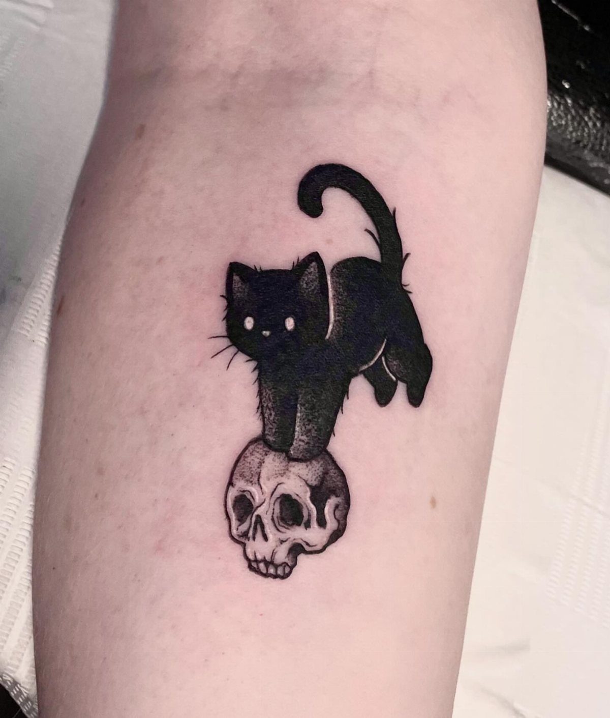 Black cat tattoo standing on a human skull on a person's arm.