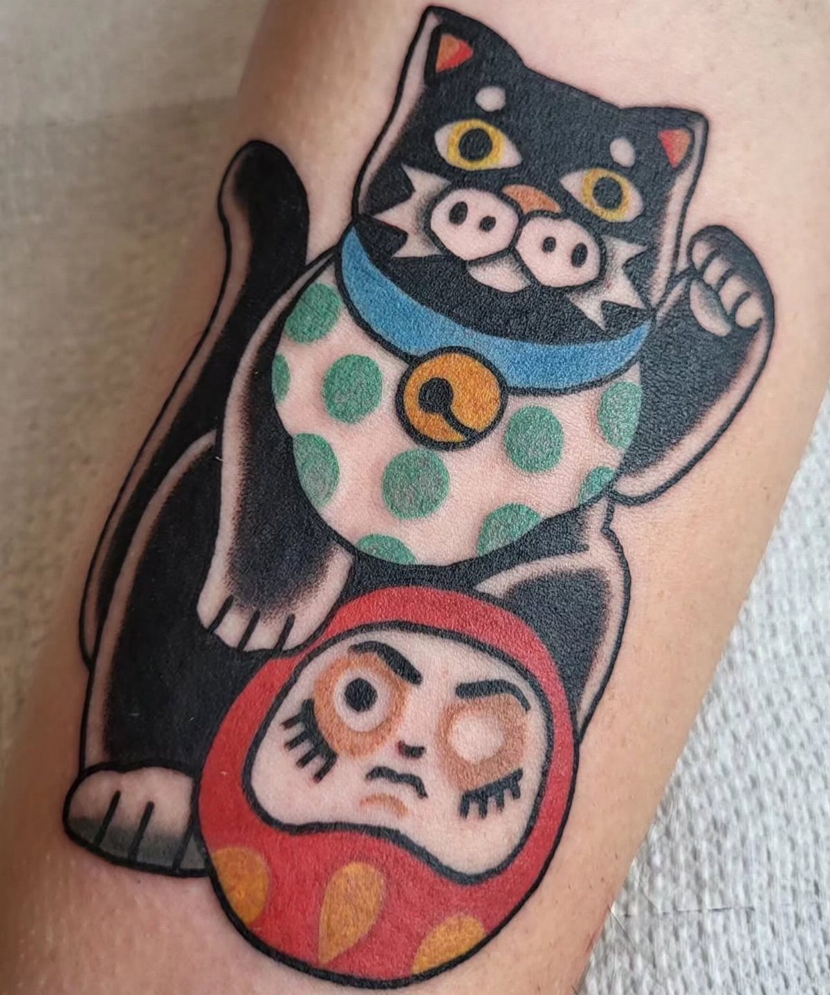 Black cat tattoo with a green polka-dot bib sitting on a red Daruma doll.
