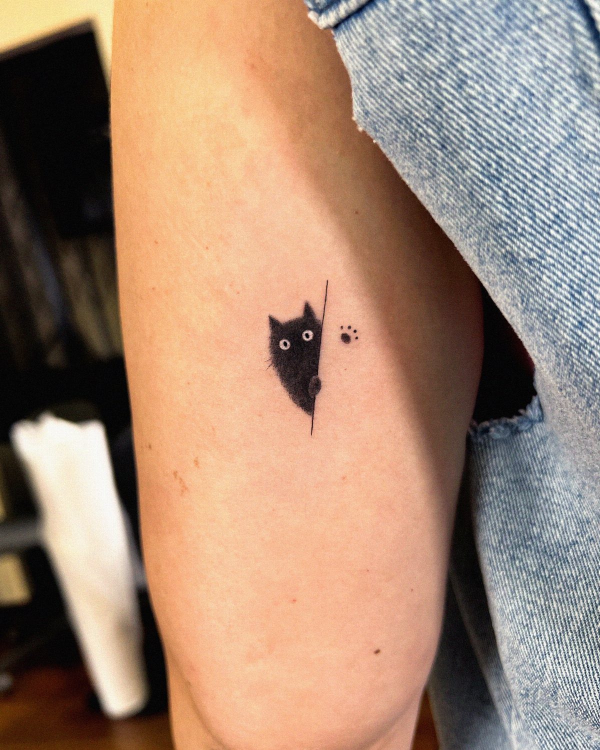 Minimalist black cat tattoo peeking from a line on a person's upper arm.