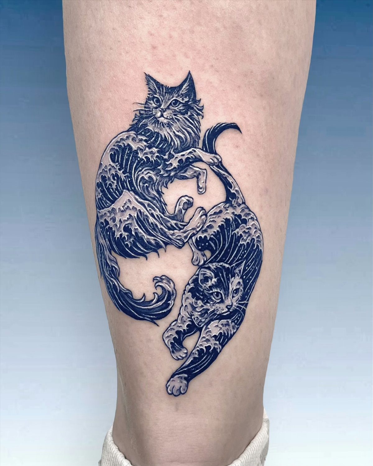 Two black cats tattooed in a yin-yang style with wave patterns on a leg.
