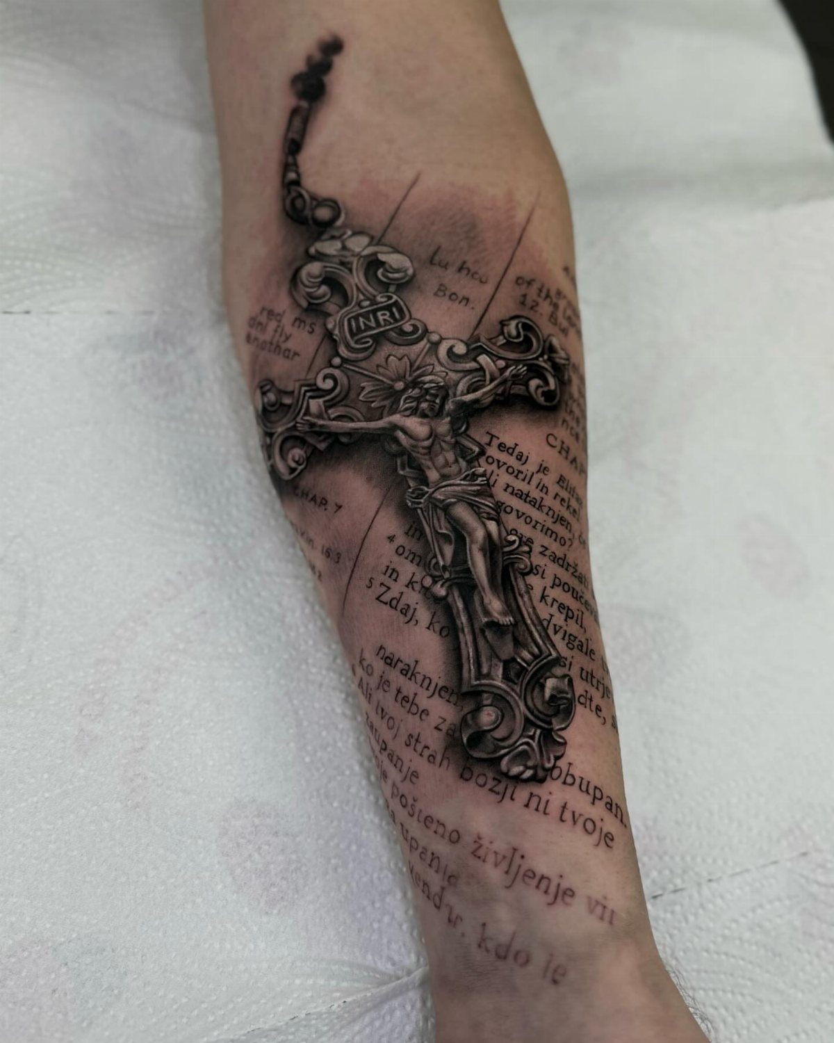 Black and grey realism tattoo of a crucifix over tattooed scripture on a forearm