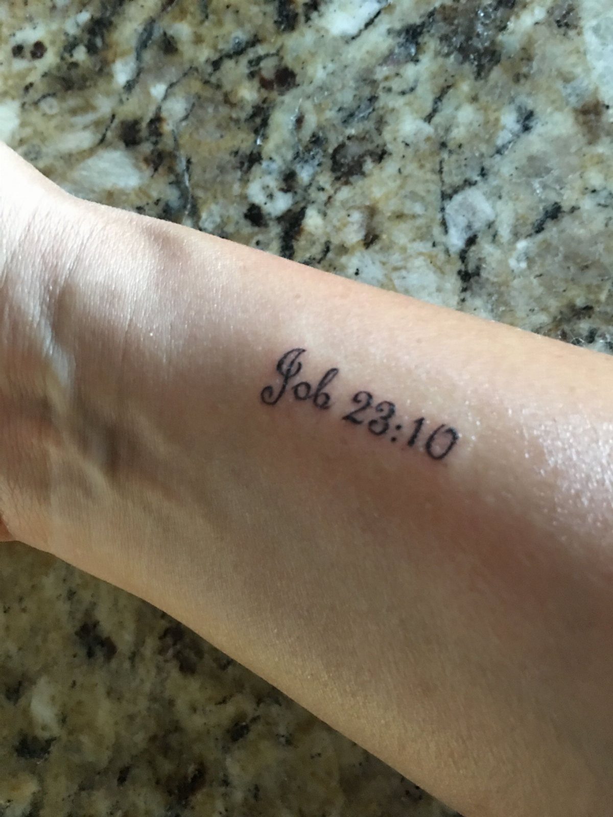 Wrist tattoo with the biblical reference Job 23:10 written in black cursive script.