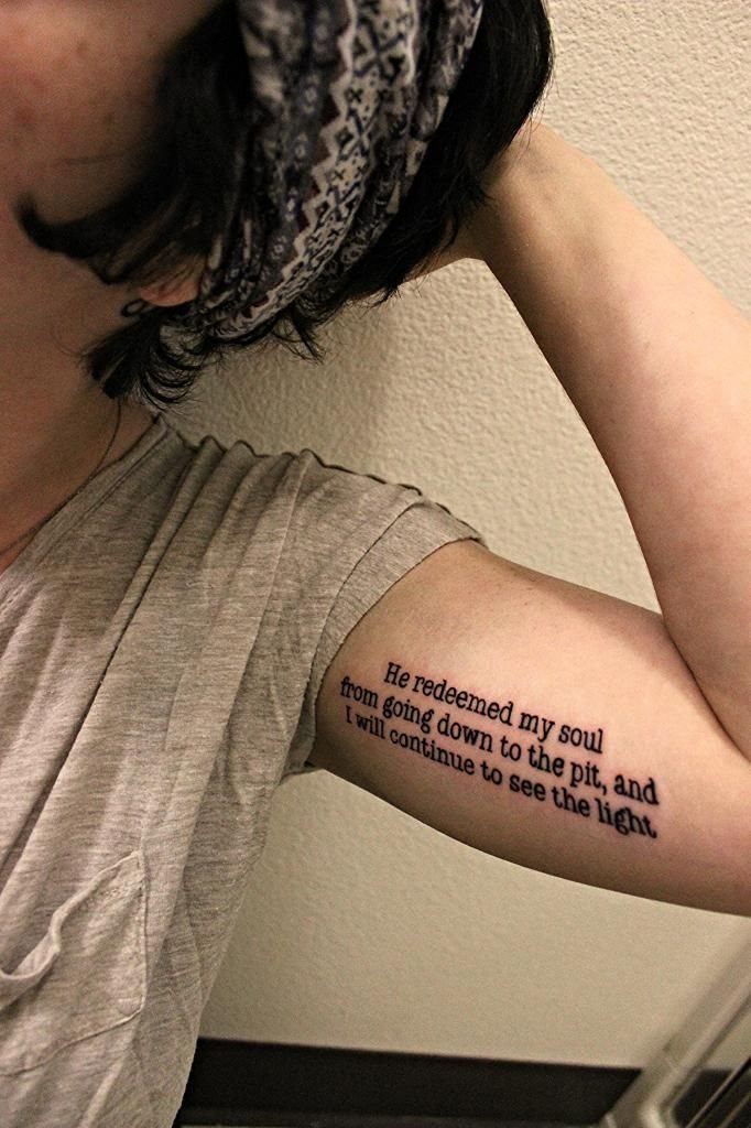 Tattoo on an upper arm with scripture text in black serif font.