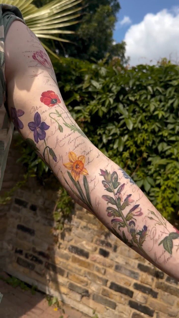 Botanical tattoo sleeve on an arm featuring colorful flowers and vintage cursive script.