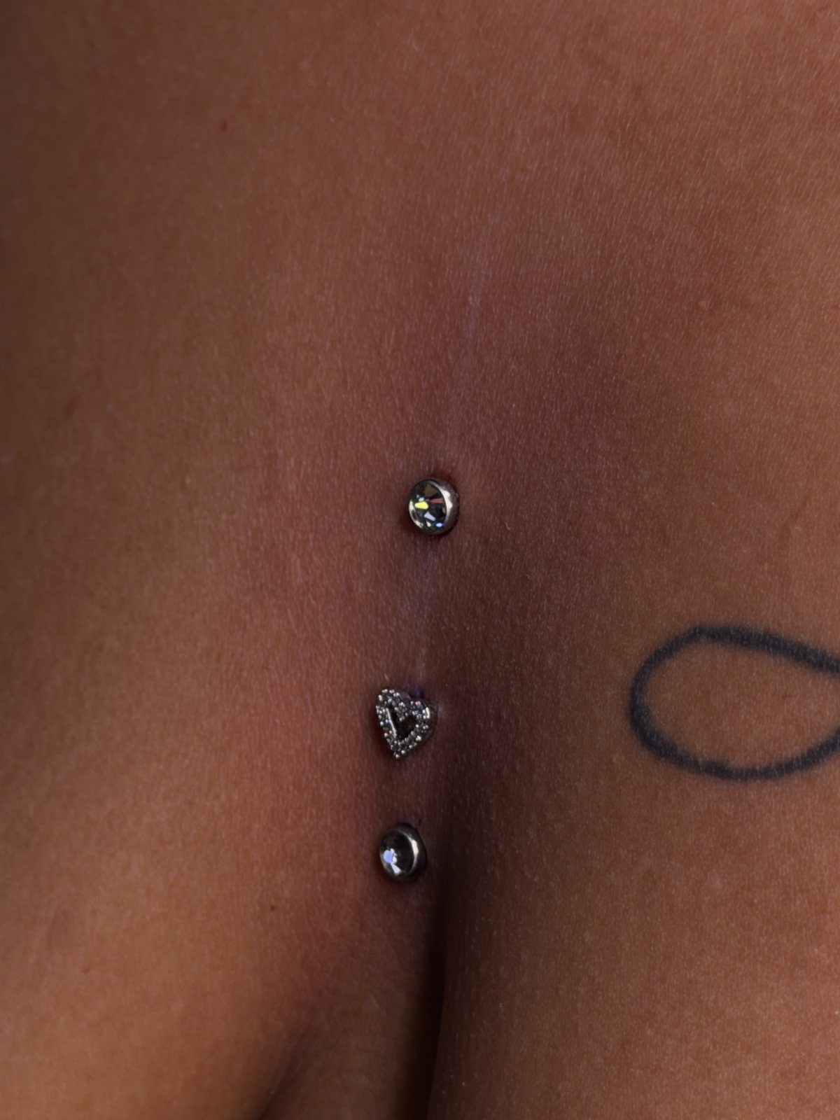 Three vertical chest piercings with clear and heart-shaped gemstones on brown skin.