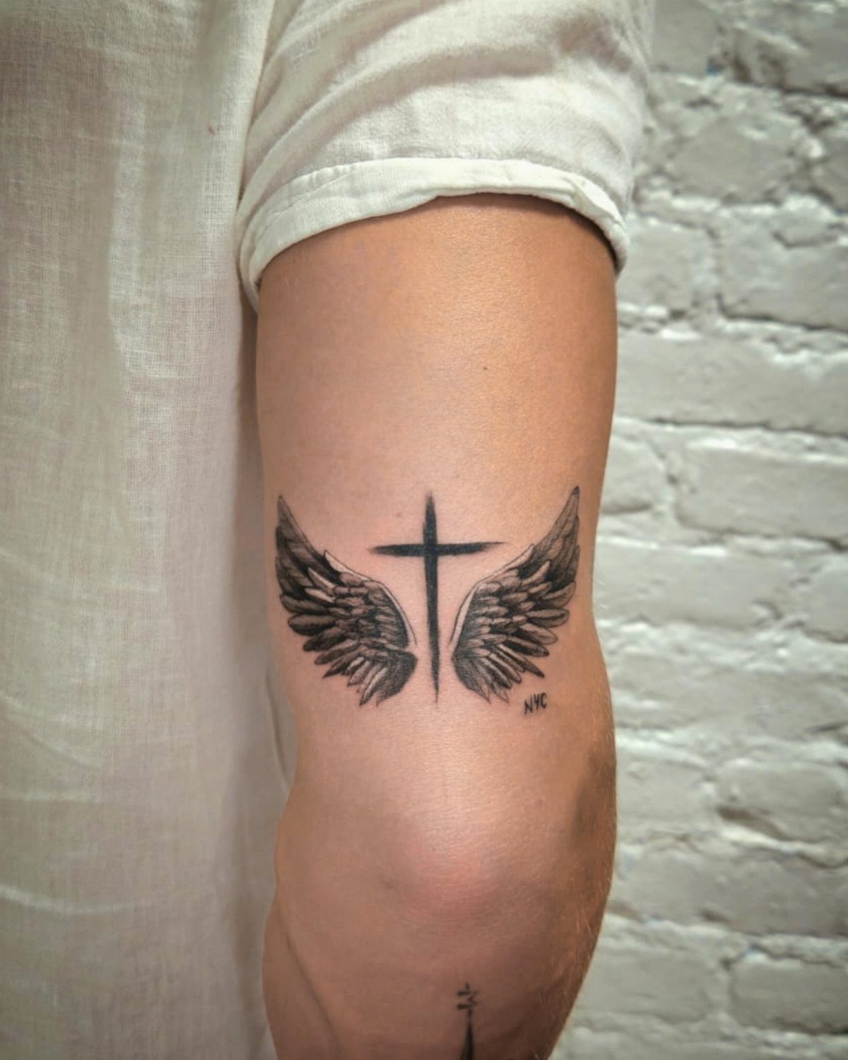 Cross tattoo with angel wings on a person's leg against a white brick wall