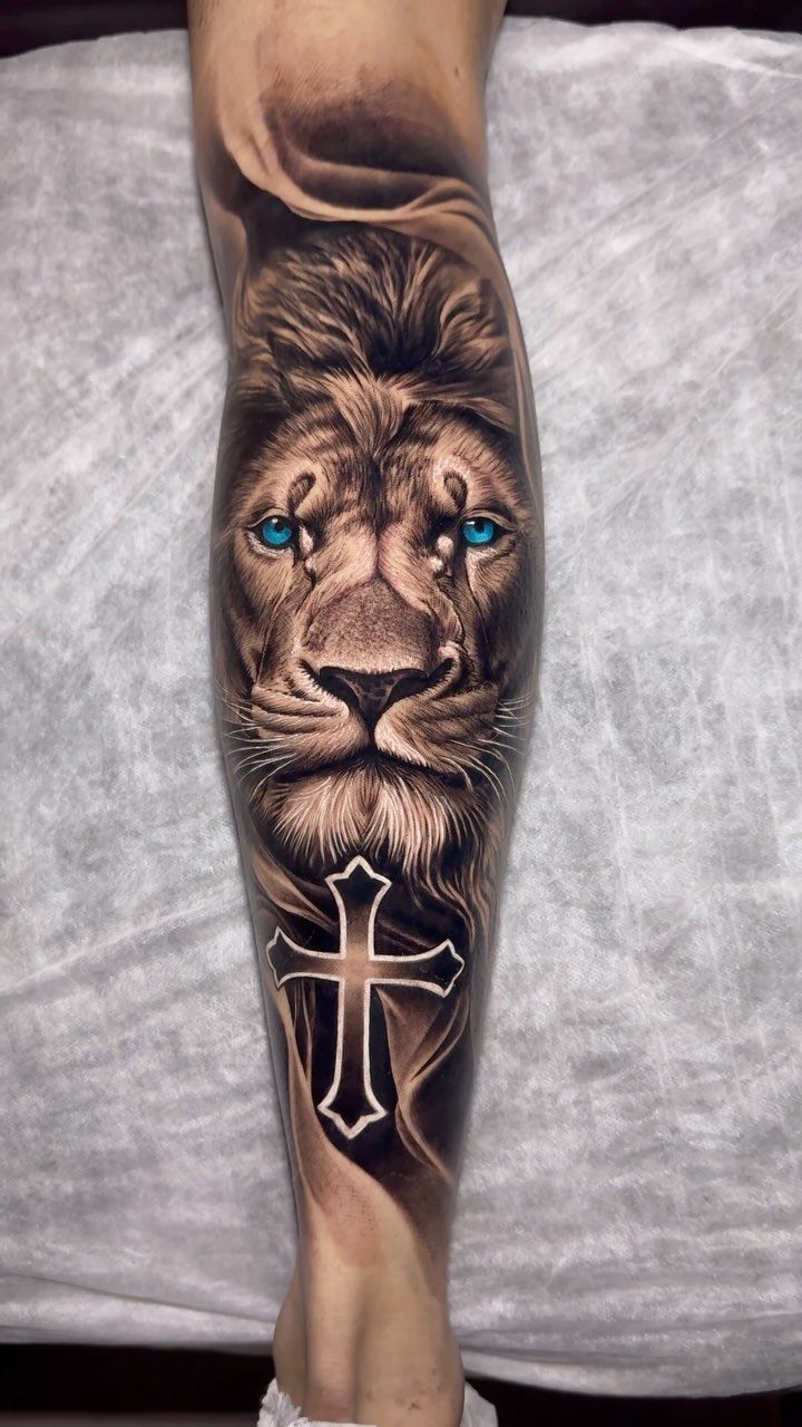 Realistic lion head tattoo with blue eyes and a cross on a leg