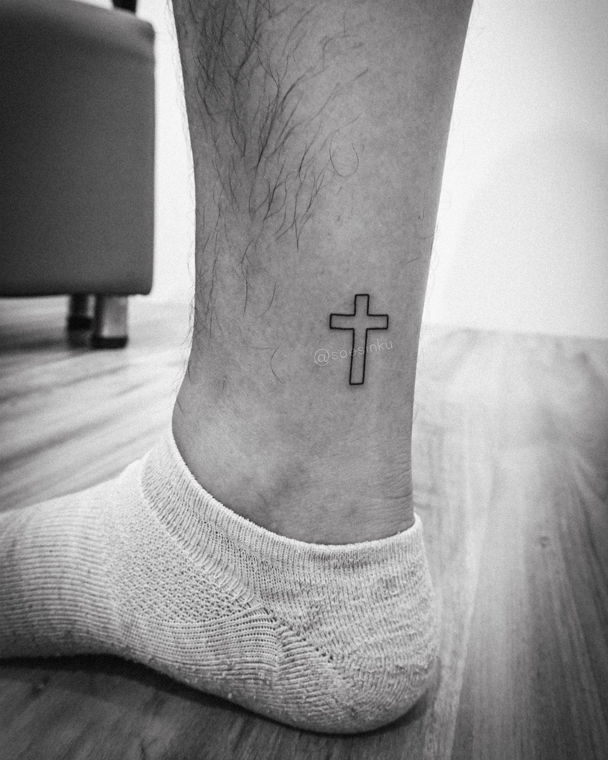 Small simple black ink cross tattoo on a person's ankle wearing a white sock.