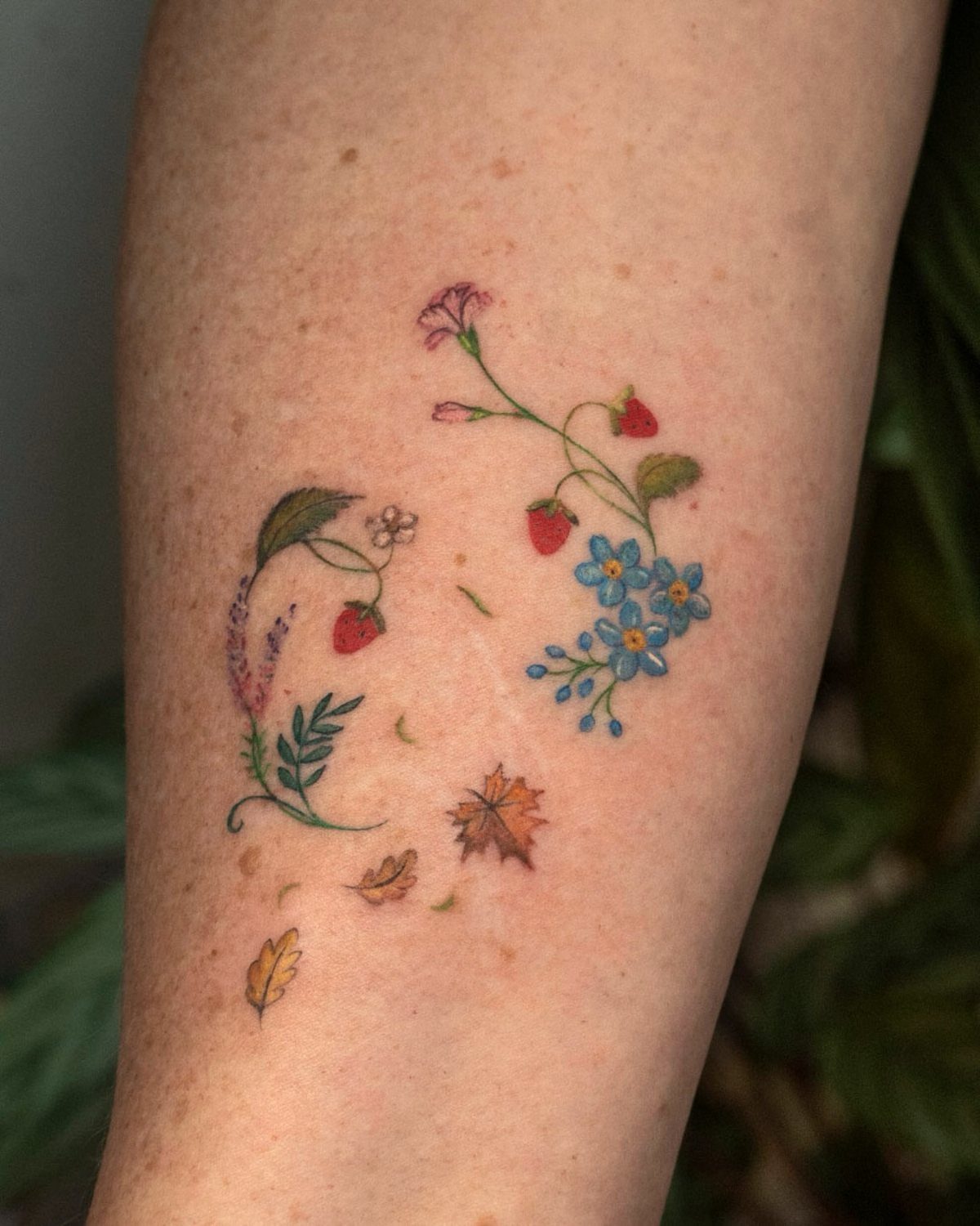 Small colorful tattoos of strawberries, blue flowers, and autumn leaves on a person's arm.