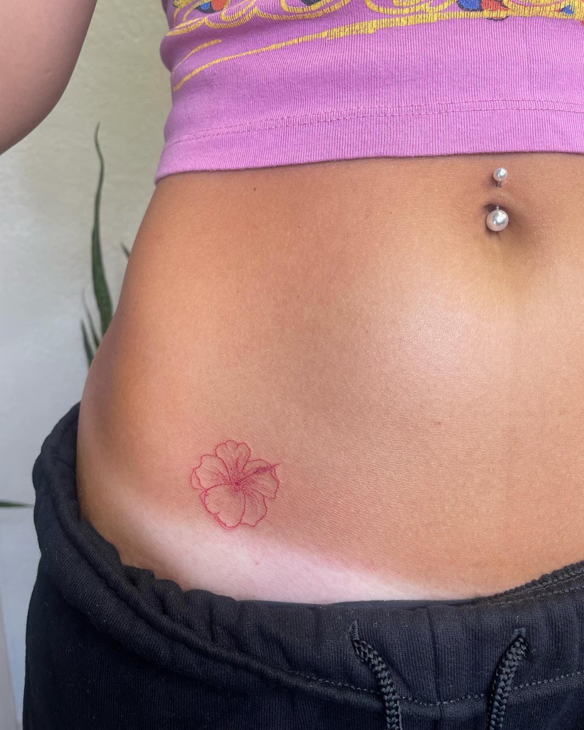 Small red hibiscus flower tattoo on a person's lower hip and midriff.