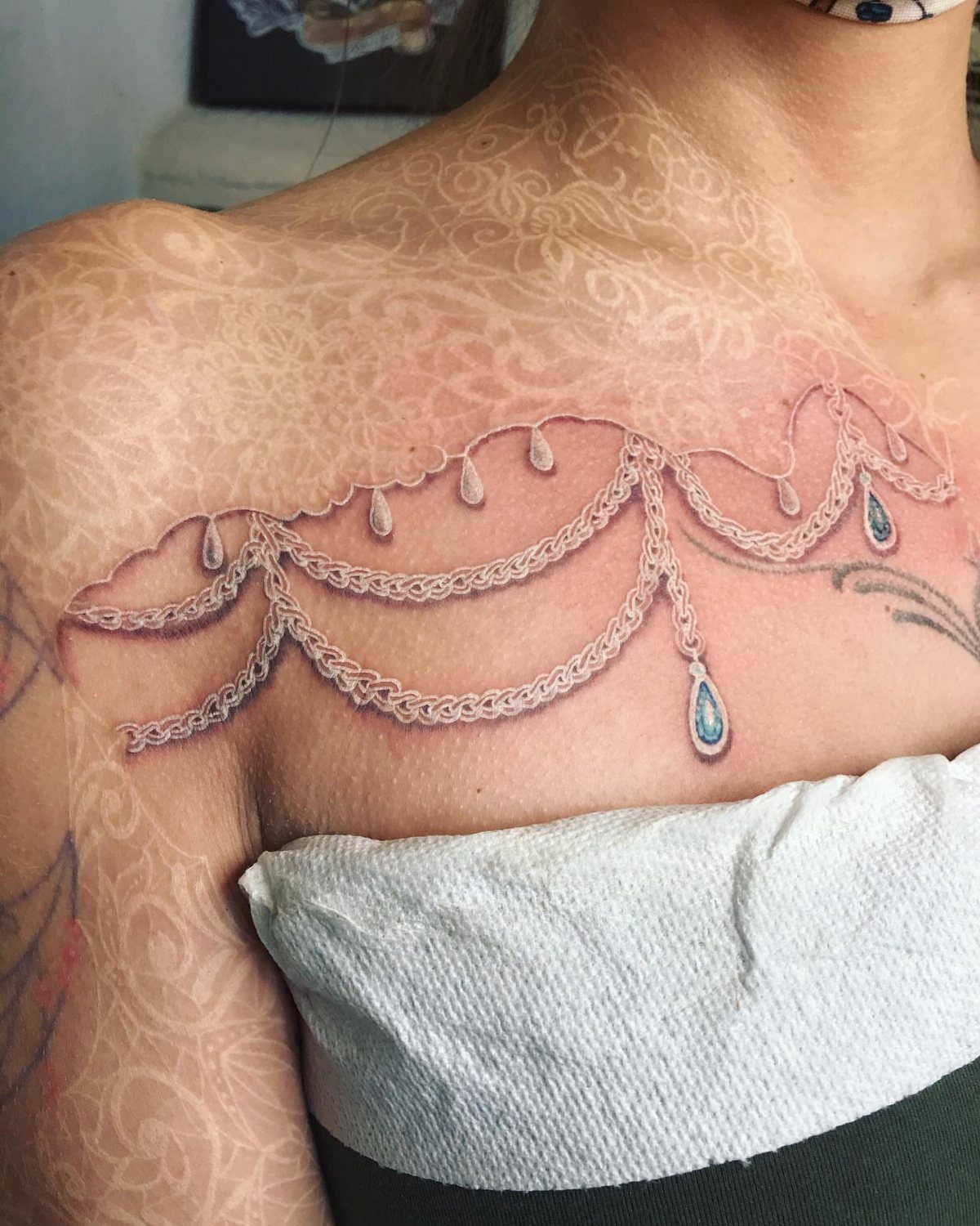 White lace and jewelry tattoo on a person's shoulder with blue gem accents.