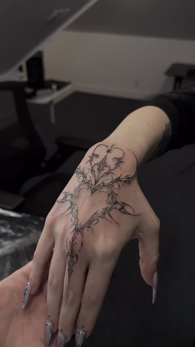 Hand with a black ink cybersigilism tattoo featuring sharp, thorny heart-shaped lines.