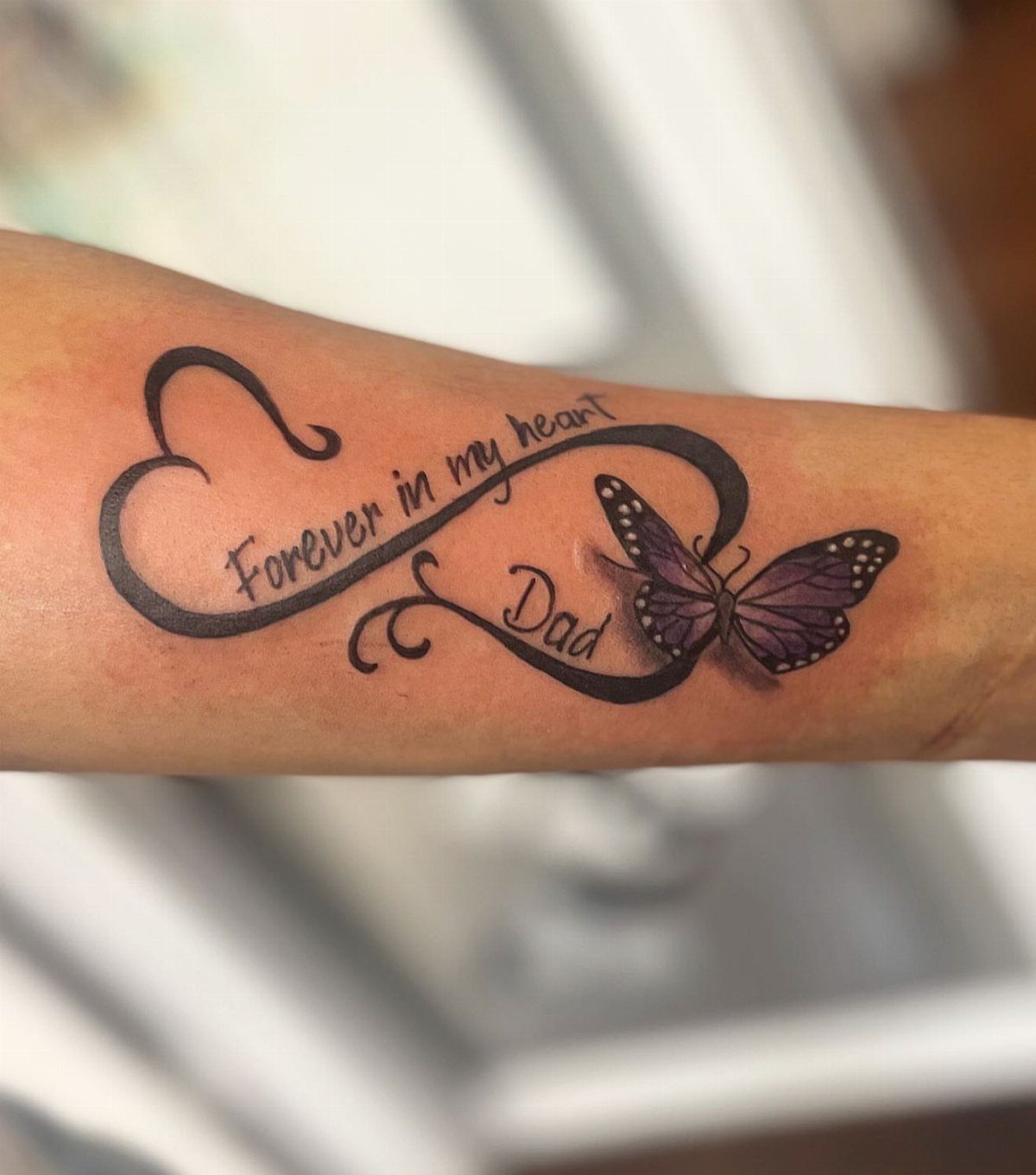 Infinity symbol tattoo with a purple butterfly and the word Dad on forearm