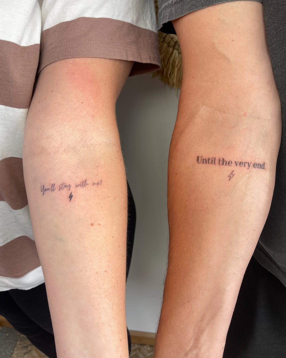 Two forearms with matching quote tattoos and small lightning bolt symbols in black ink.