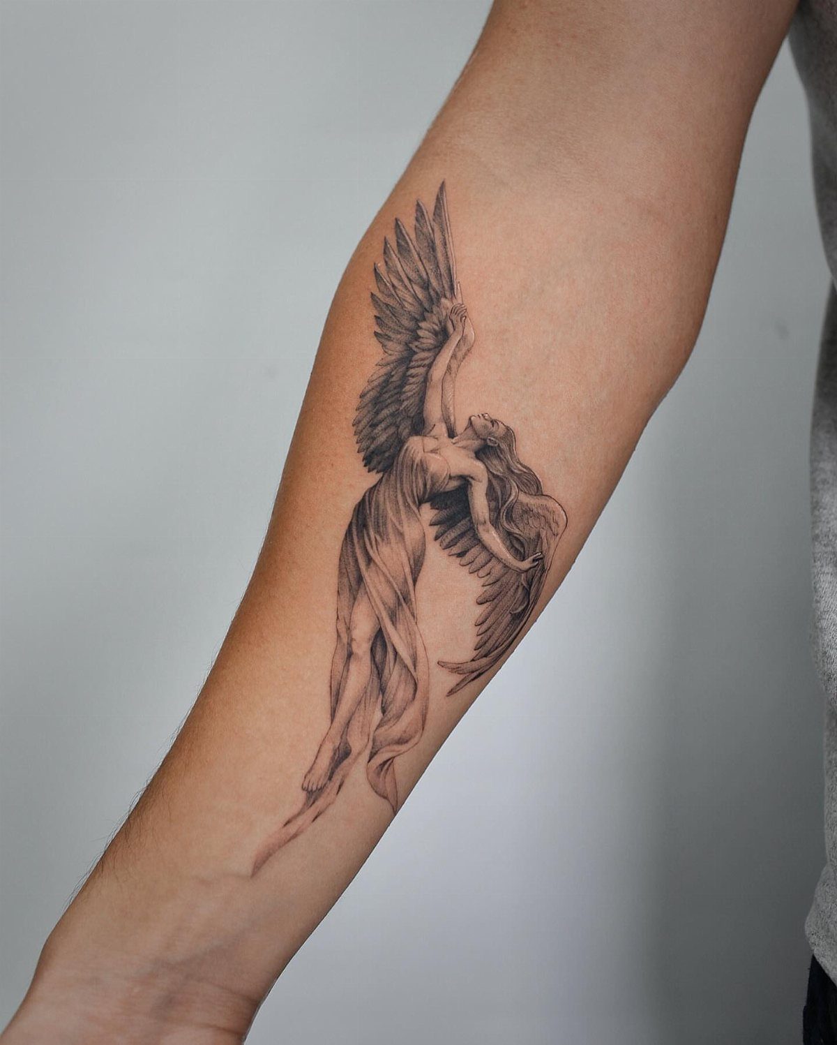 Winged female angel tattoo with flowing robes on a person's forearm.