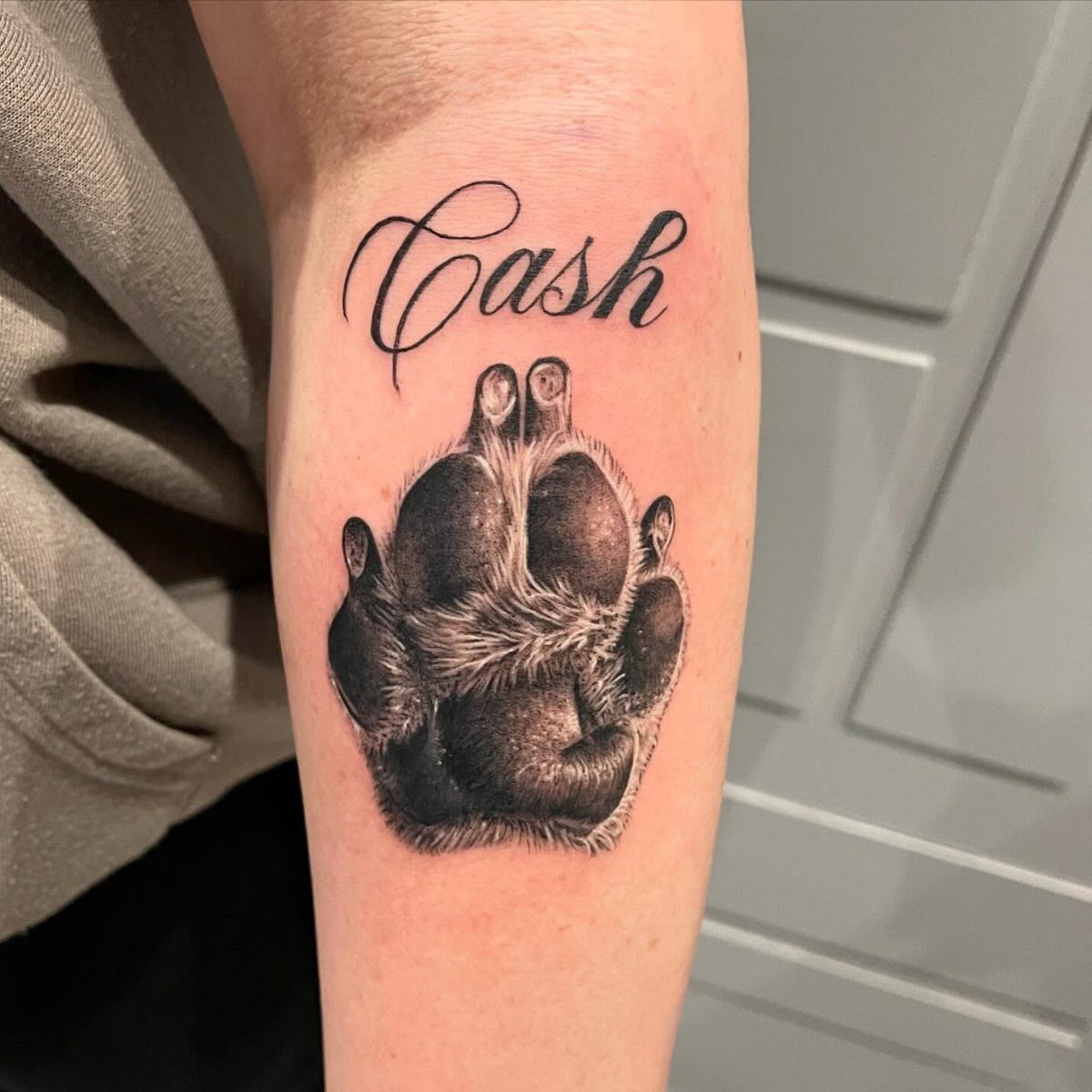 Dog paw print tattoo with the name Cash written in cursive on a forearm