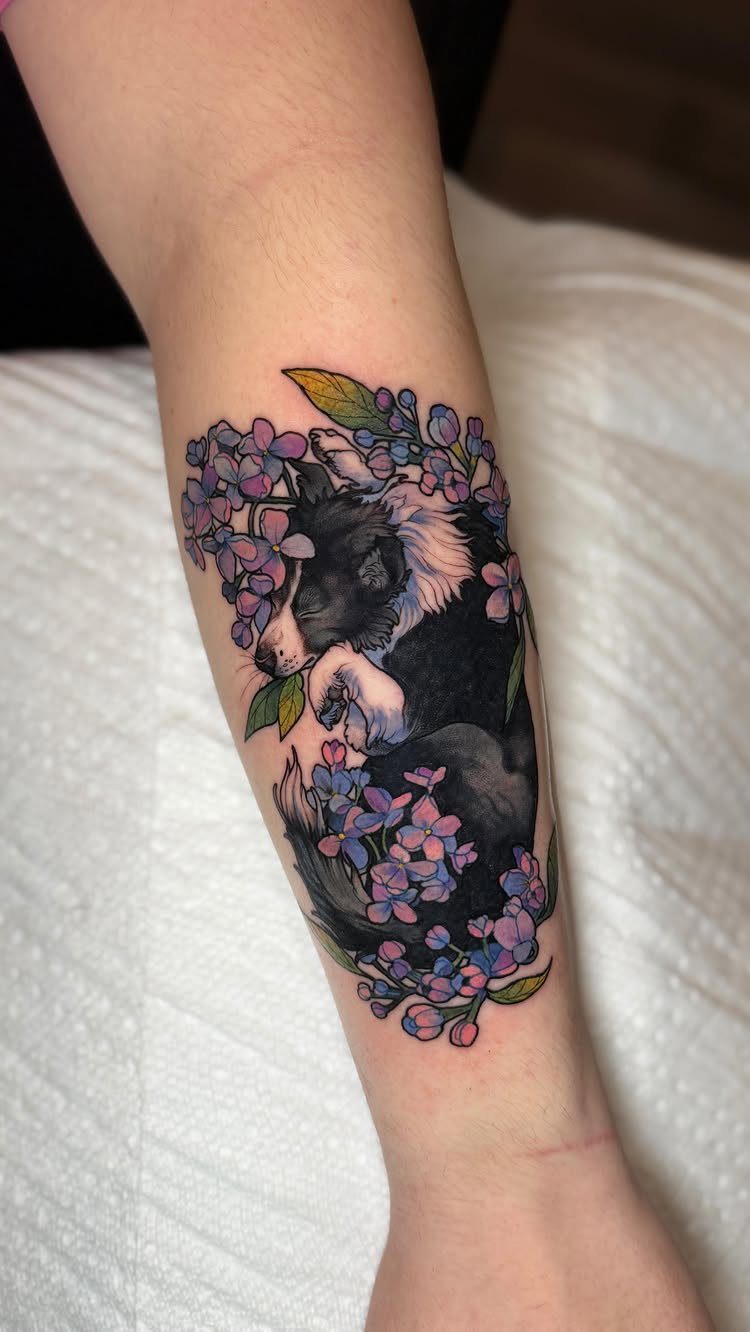 Dog tattoo on a forearm surrounded by purple and blue flowers.
