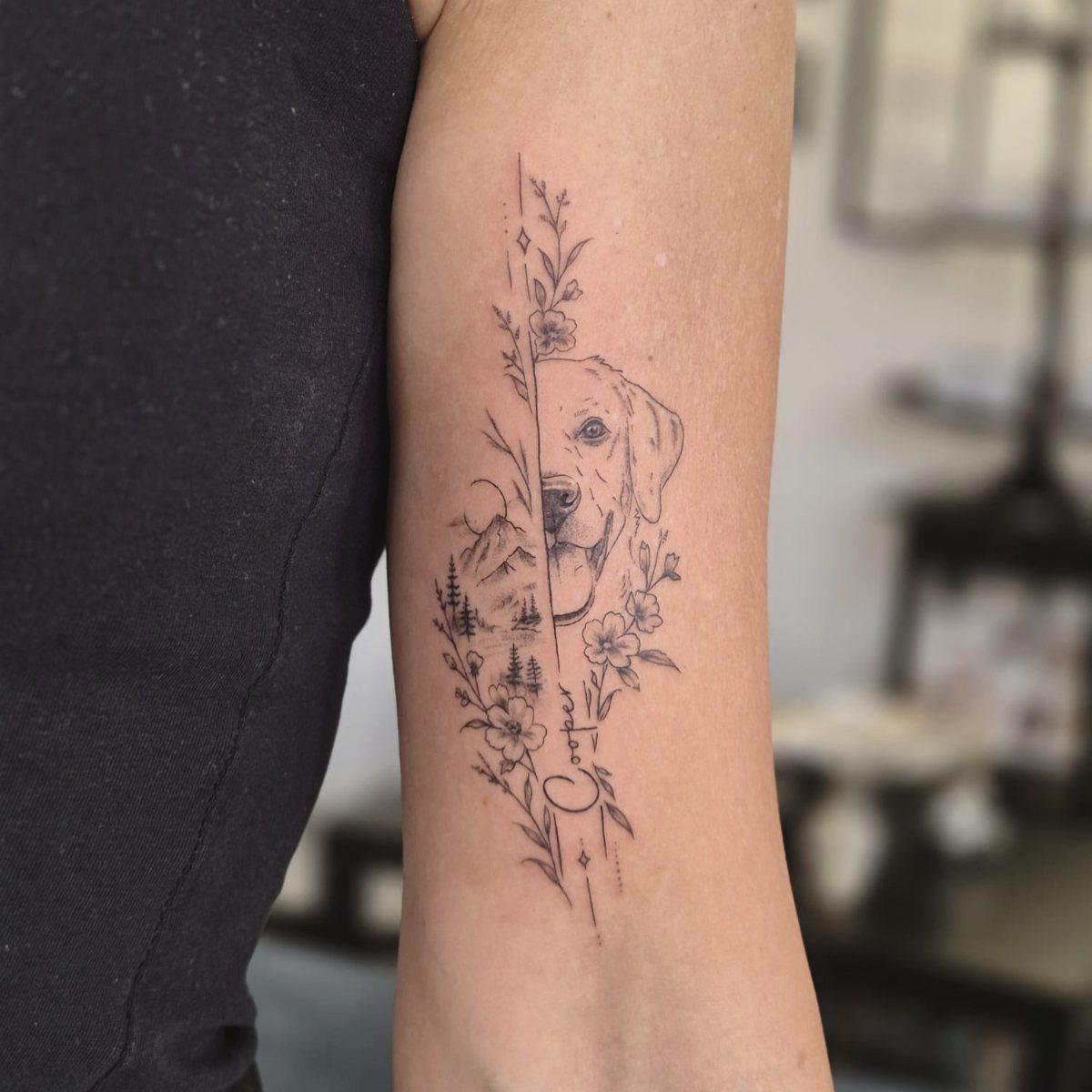 Dog portrait tattoo merged with a mountain landscape and flowers on a forearm.