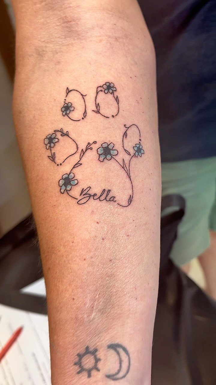 Dog paw print tattoo with blue flowers and the name Bella on a forearm.