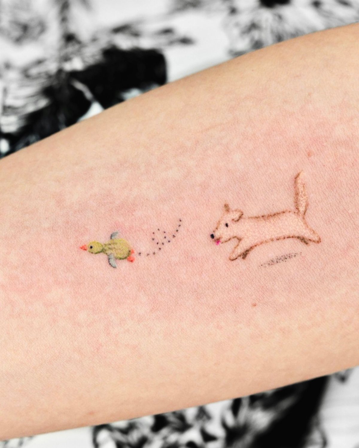 Small colorful tattoo of a brown dog chasing a yellow bird on skin