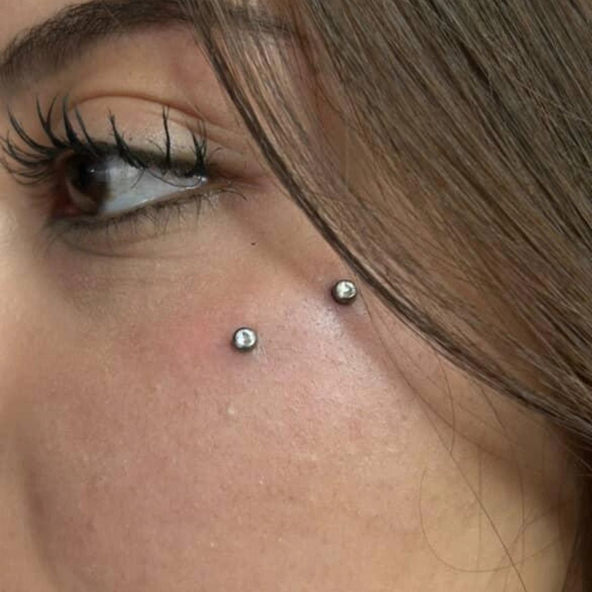 Two silver stud piercings on a person's cheek near the eye and cheekbone.