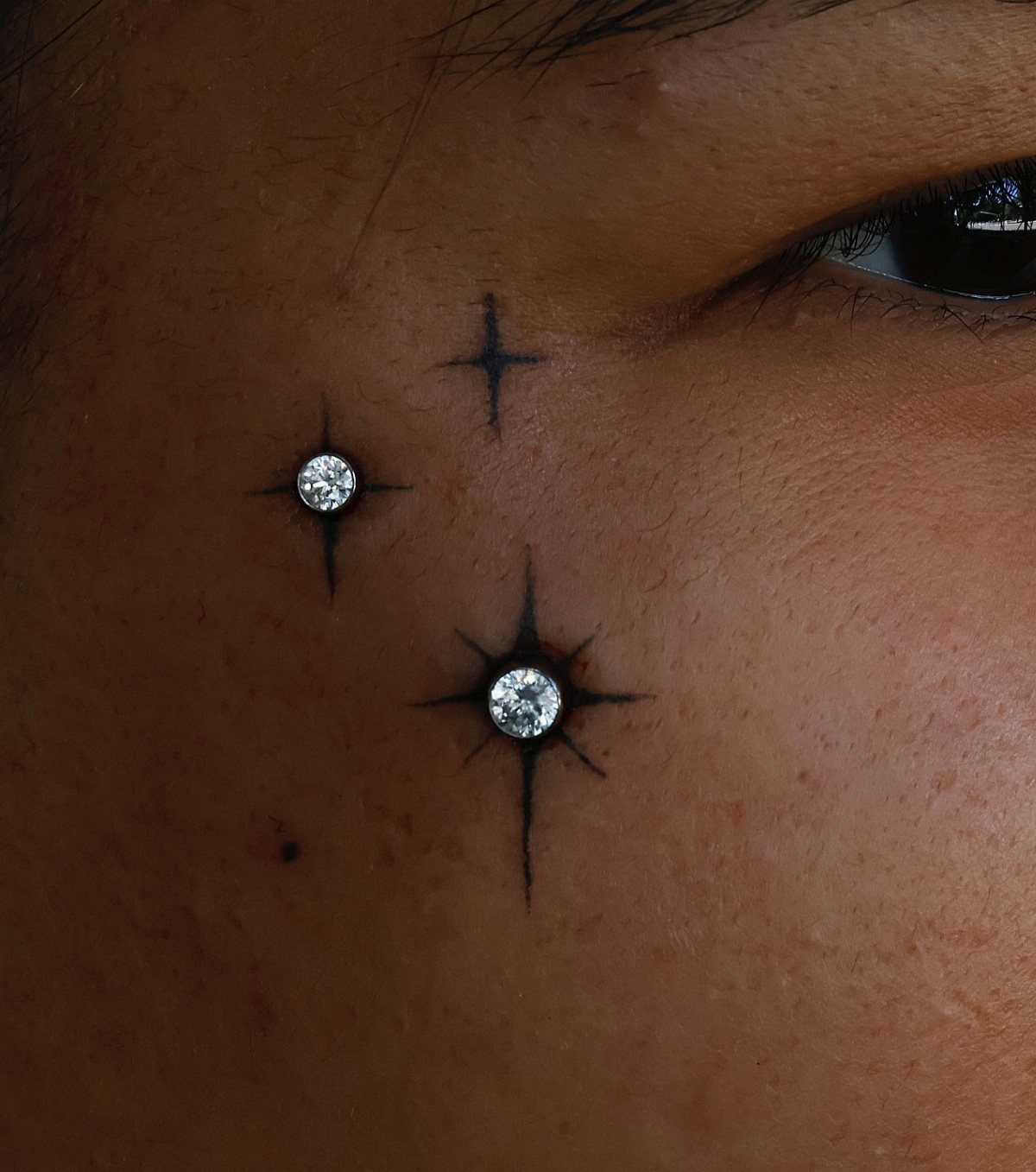 Cheekbone piercings with diamond studs and black star tattoos on a person's face.
