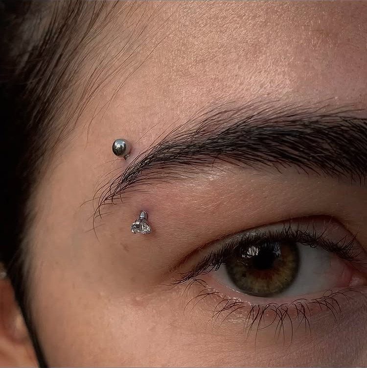 Person's eyebrow and eye featuring a silver ball and gemstone piercing.