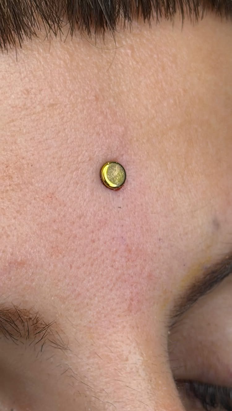 Gold circular stud piercing centered on a person's forehead between the eyebrows.