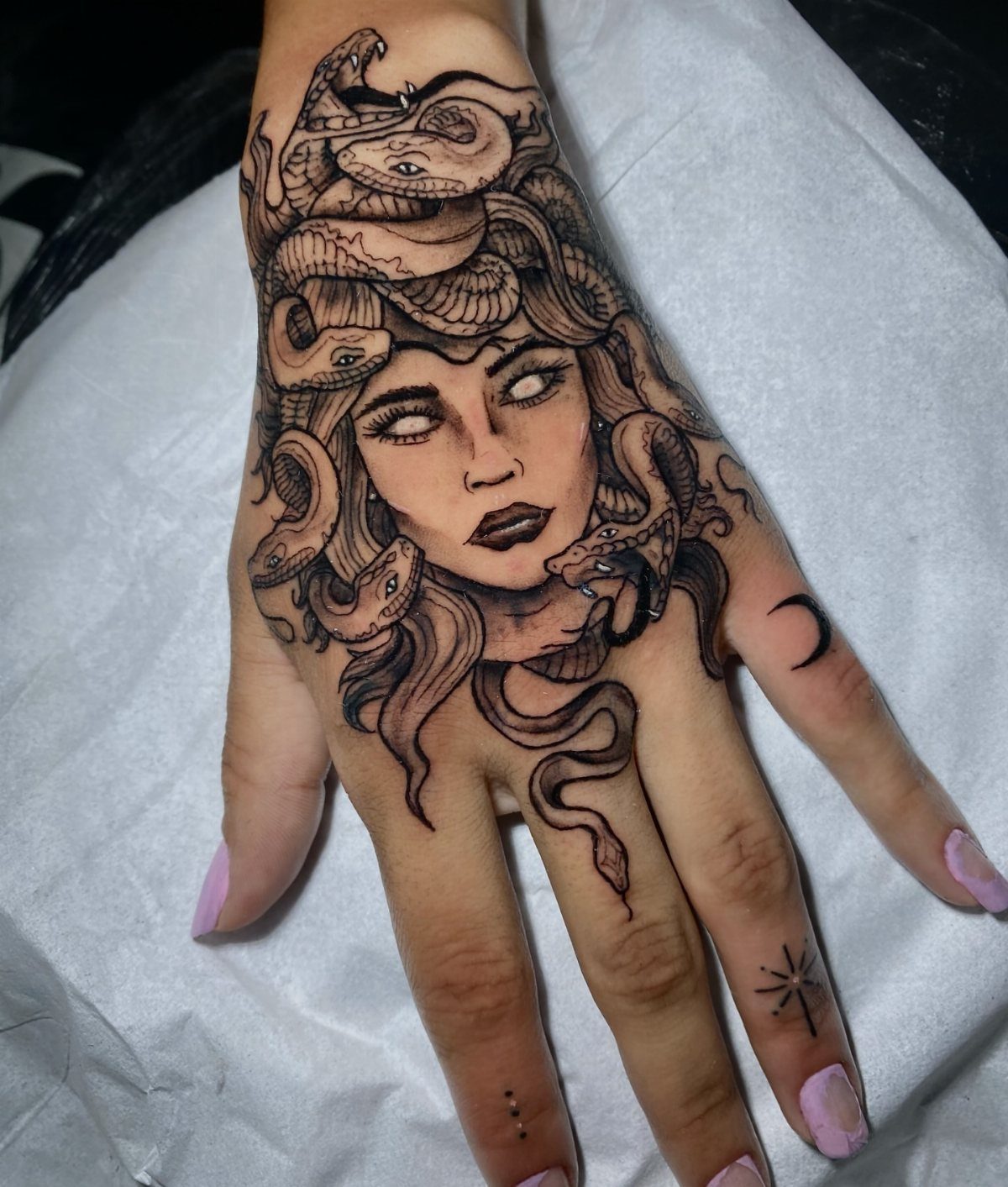 Medusa tattoo on a hand with snakes for hair and blank white eyes.