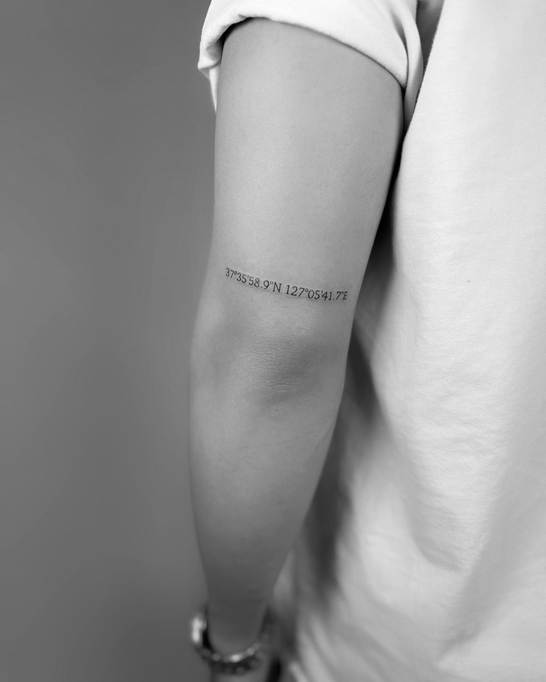 Forearm tattoo of geographic coordinates in a clean, minimalist black ink font.