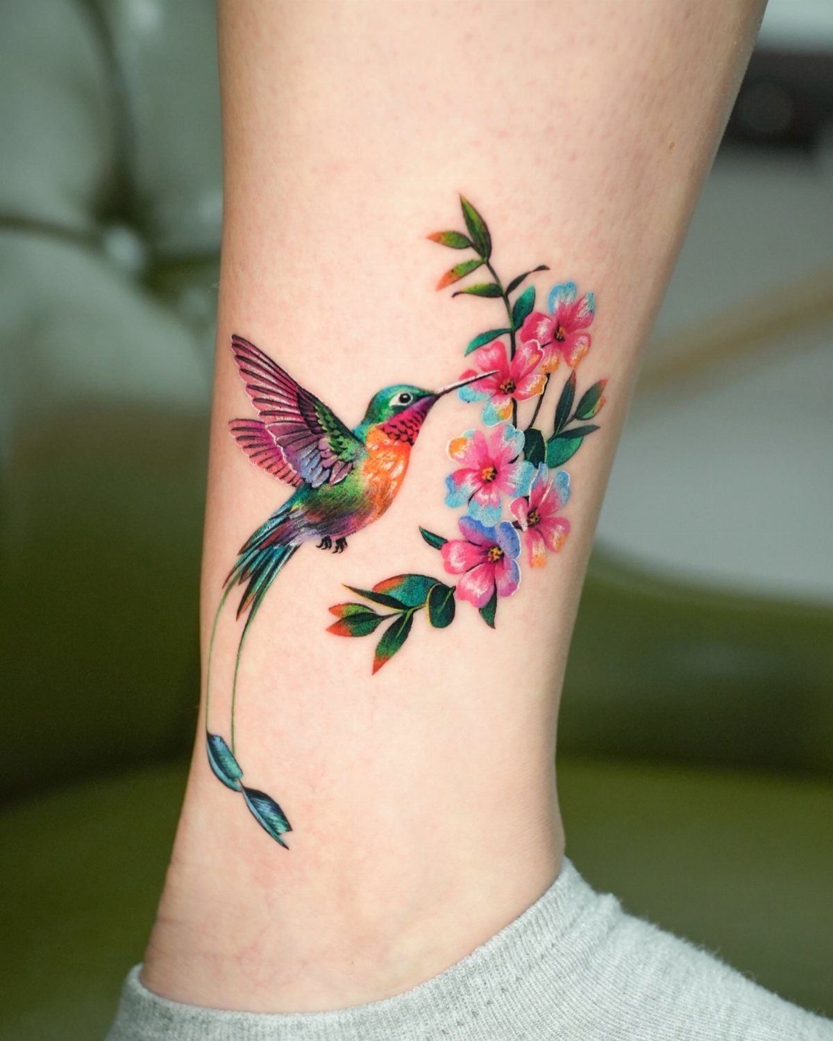 Colorful hummingbird tattoo with pink flowers on a person's ankle.